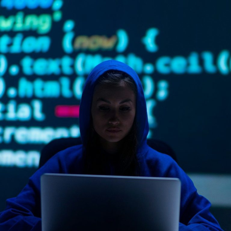 Person in Kapuzenpullover sitzt vor Laptop, Code im Hintergrund leuchtet.