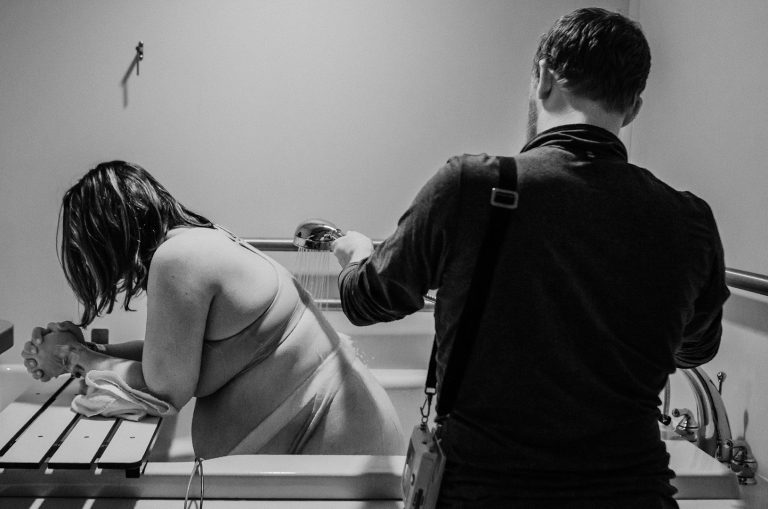 Eine Person steht neben einer anderen, die in einer Badewanne sitzt. Schwarz-weiß Fotografie.