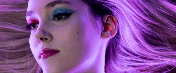 Frau mit buntem Augen-Make-up und langen, fließenden Haaren vor einem lila Hintergrund.