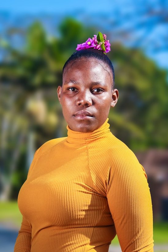 Porträt einer Frau mit orangefarbenem Oberteil und lila Haaraccessoire vor einer grünen Kulisse.